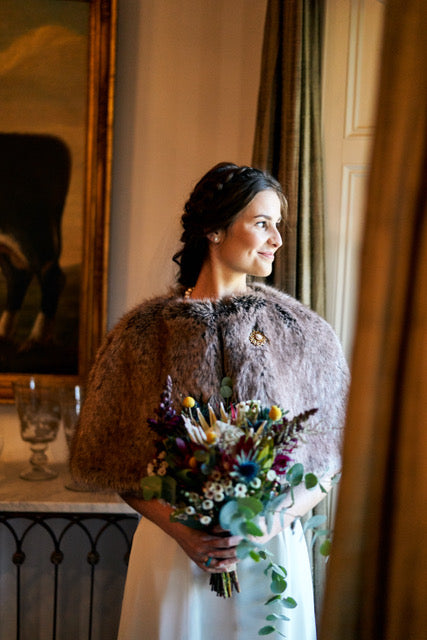 Bride standing with a bouquet wearing a light brown faux fur short cape by Helen Moore